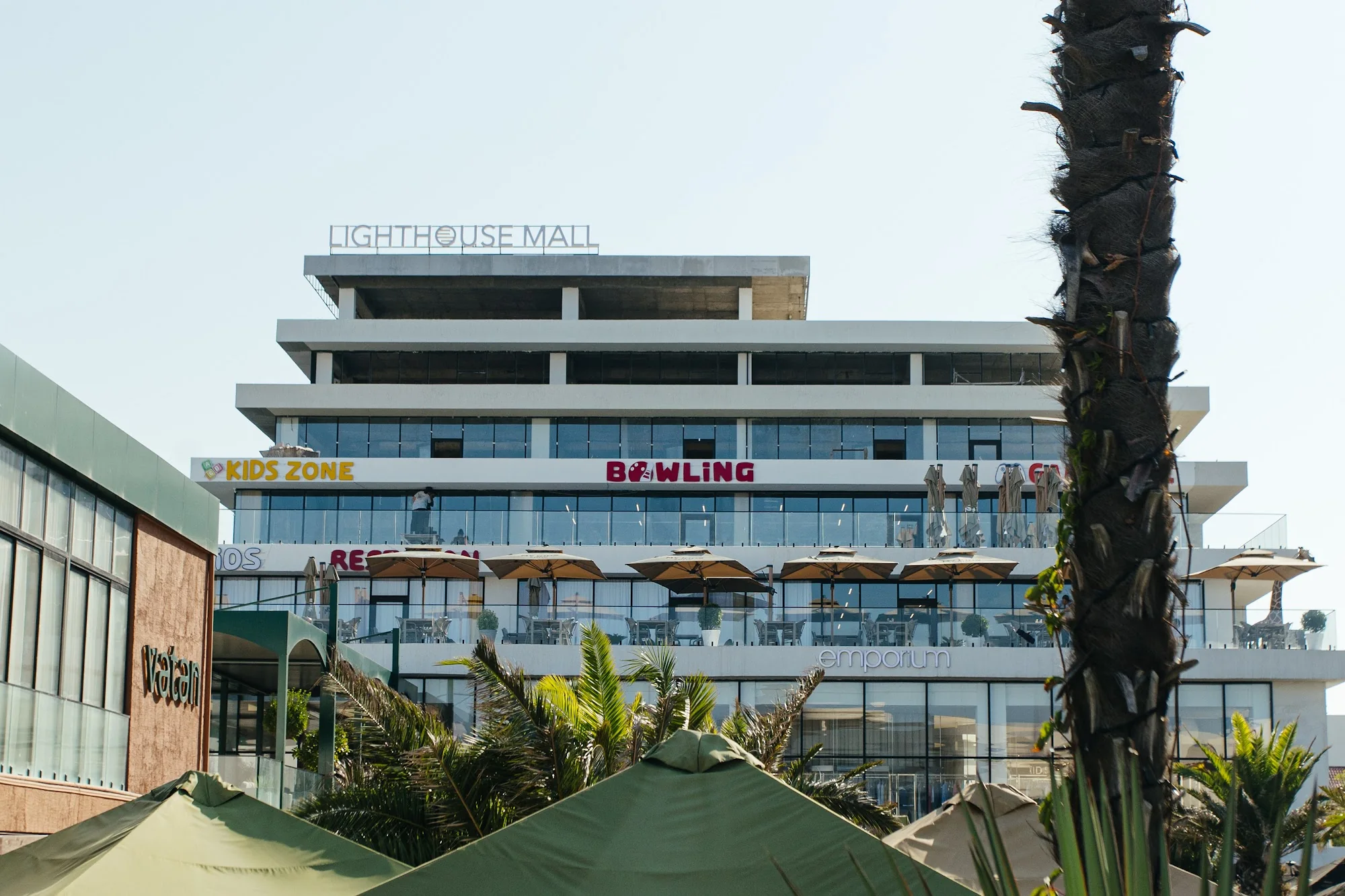 Lighthouse Mall — ступенчатое здание с вывесками KIDS ZONE и BOWLING на верхнем ярусе, ниже терраса с зонтами и Emporium. Салон 15/25 Studio — в этом здании; его вывески на снимке не видно